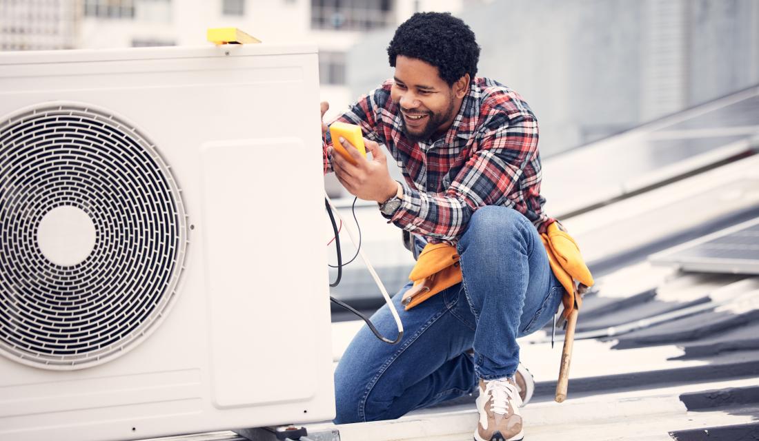 Ein freundlicher Techniker sitzt mit einem Testgerät neben einer elektrischen Wärmepumpe.