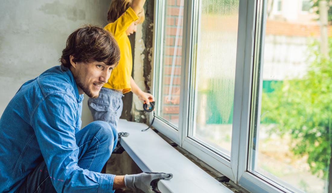 Ein Mann tauscht ein Fenster aus. Er montiert gerade die Fensterbank an oder ab. Ein kleiner Junge unterstützt ihn.